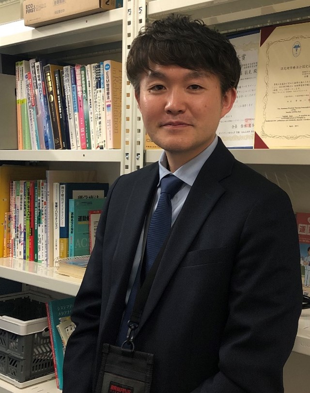 福島県立医科大学に着任した佐藤聡見さんが書棚の前で微笑んでいる写真。