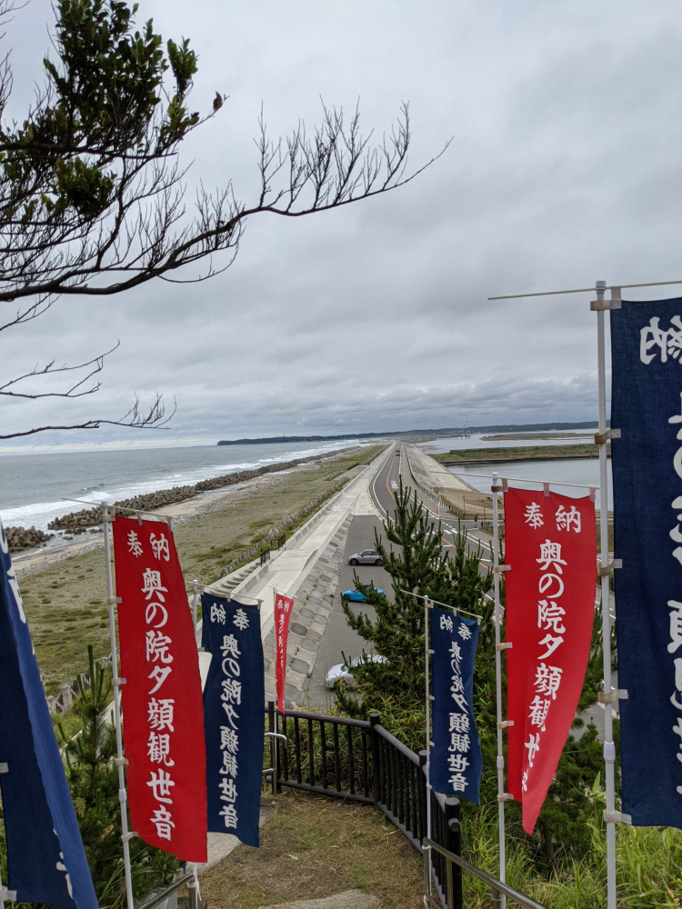 松川浦を見渡す堤防の風景と、復興を願う旗が立つ様子。曇り空の下、海岸線が続いている。