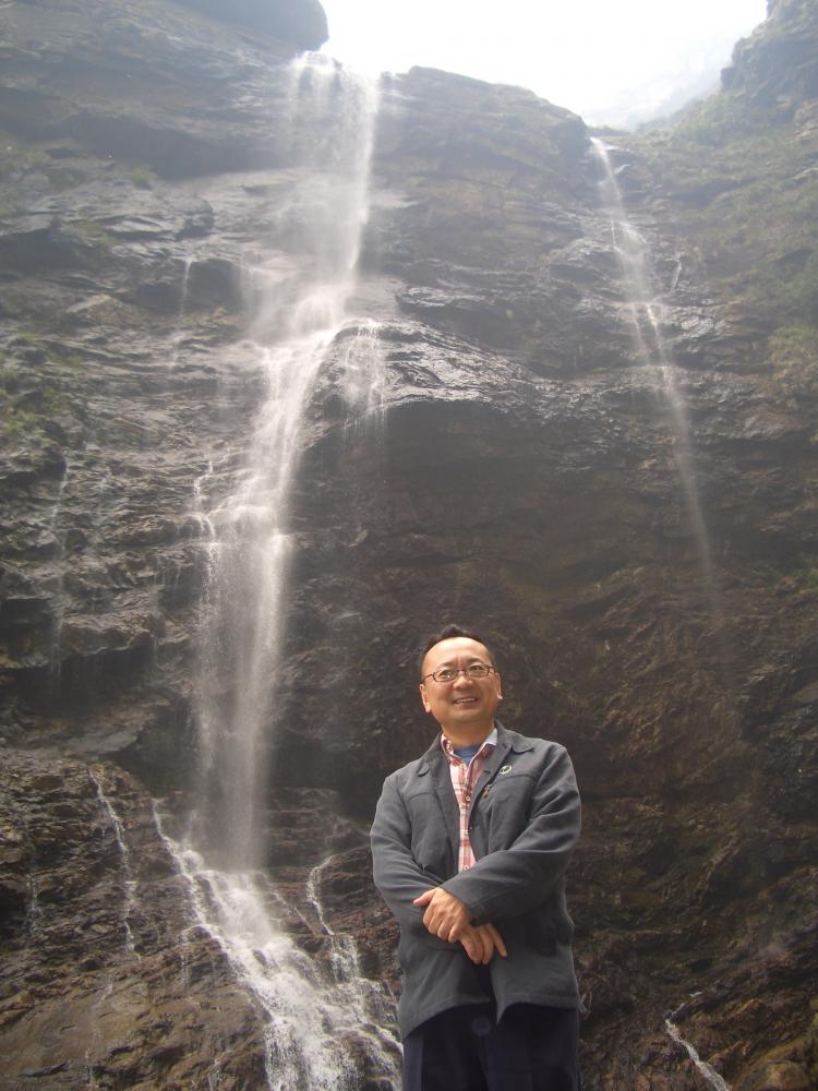 廬山の滝の前で微笑む男性の写真。滝が岩肌を流れ落ちる美しい風景が広がっている。