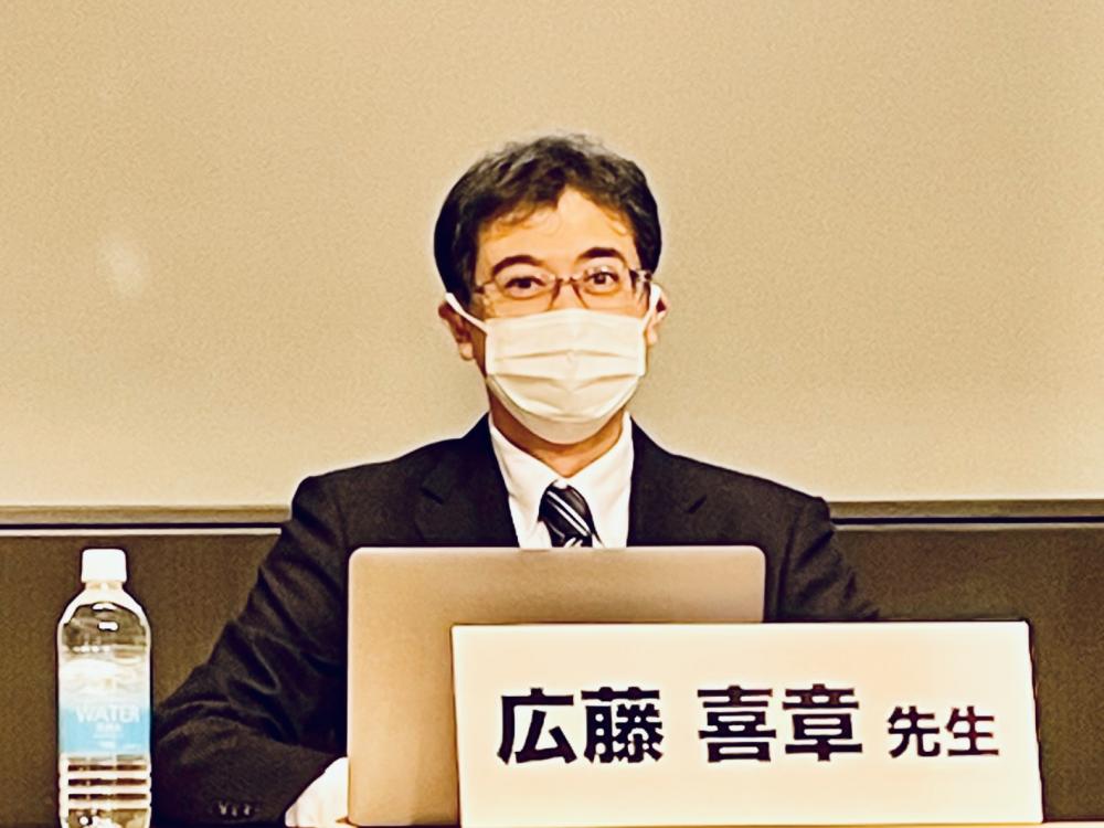 広藤喜章先生がマスクを着用し、講演中の様子。テーブルには水のボトルとパソコンが置かれている。