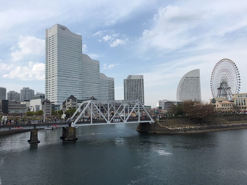 みなとみらいの遠景、近代的なビル群と観覧車が見える風景。橋が水面に架かり、賑わう人々の姿も確認できる。