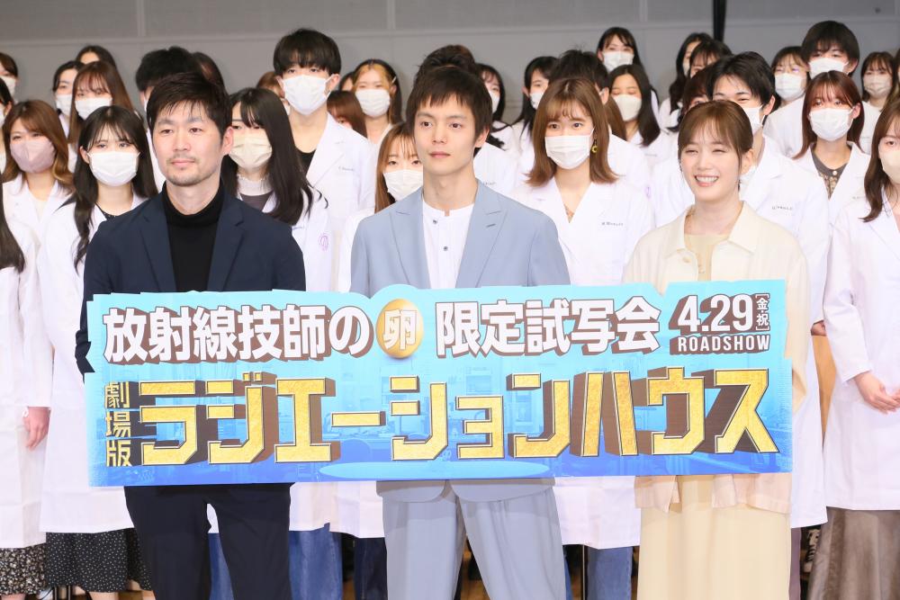 劇場版「ラジエーションハウス」の限定試写会で、白衣を着た出演者たちが集まり、映画の宣伝看板を持っている様子。