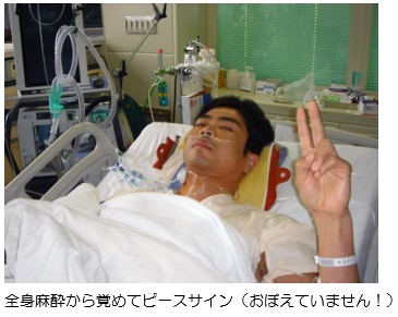 全身麻酔から覚醒した男性が病院のベッドでピースサインをしている様子。