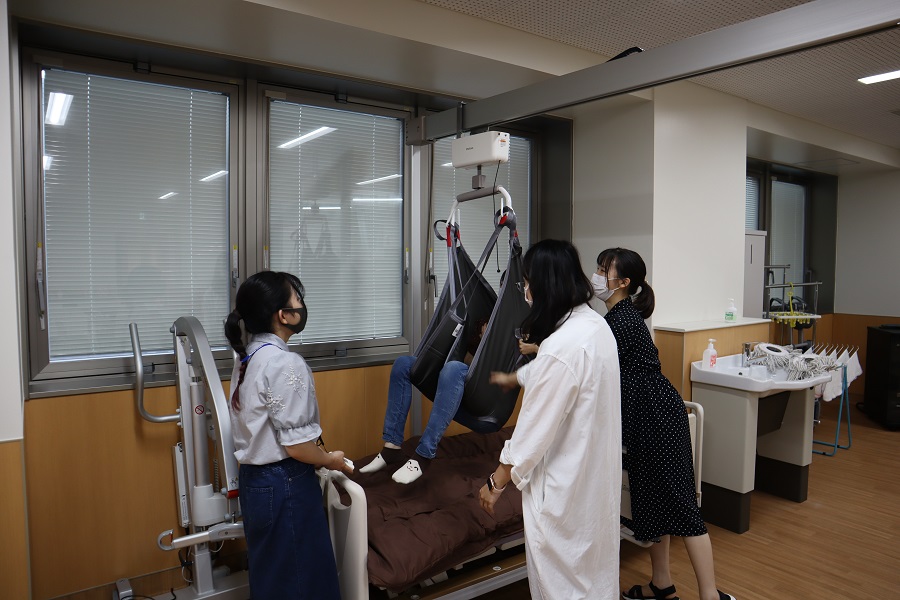 福祉用具の体験会で、利用者がリフトを使って移動する様子を参加者が見守る場面。