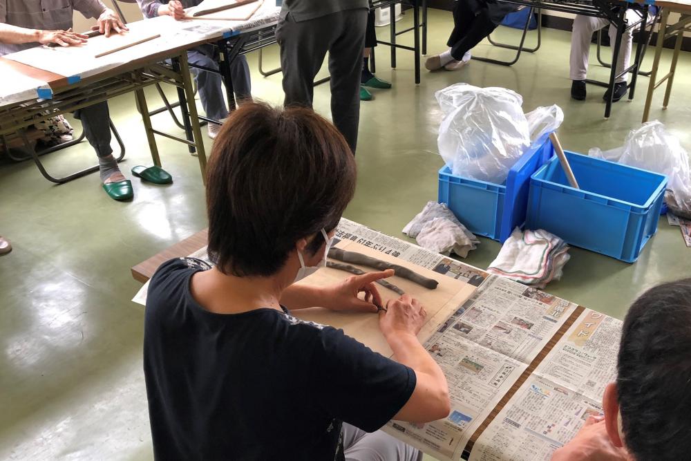 健康相談会で参加者が作業を行っている様子。テーブルには新聞紙が敷かれ、周囲には他の参加者が座っている。