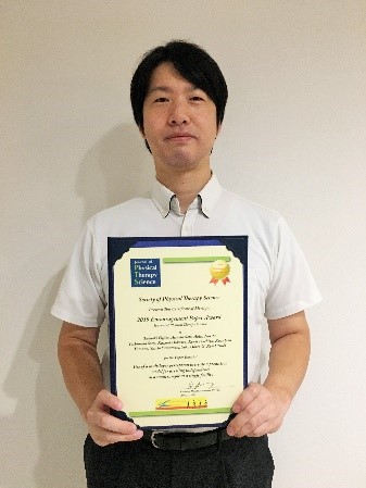 新医療系学部設置準備室の教員が学会表彰を受け、賞状を持って微笑んでいる様子。
