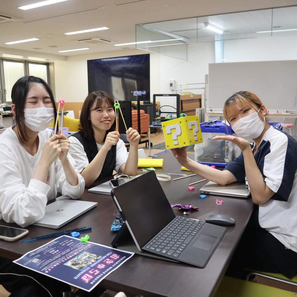 3人の学生がテーブルで楽しそうにポーズをとり、3Dプリンターコースの活動を示すアイテムを持っています。
