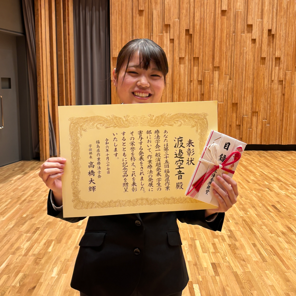 作業療法学科の学生が学生優秀演題賞を受賞し、賞状を持って笑顔でポーズをとっている。背景には木目調の壁が見える。