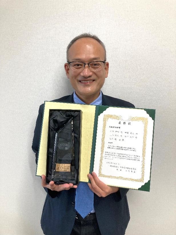 作業療法学科の倉澤教授が最優秀演題賞を受賞し、賞状とトロフィーを持って笑顔でポーズをとっています。