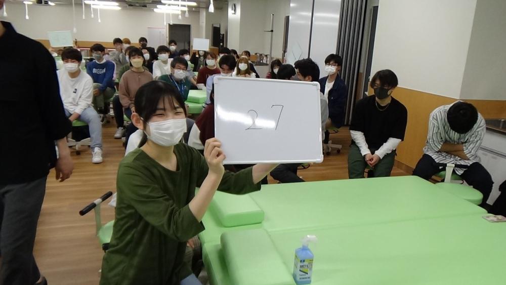 学科交流会で「27」と書かれたホワイトボードを持つ女性と、参加者たちが座っている様子。