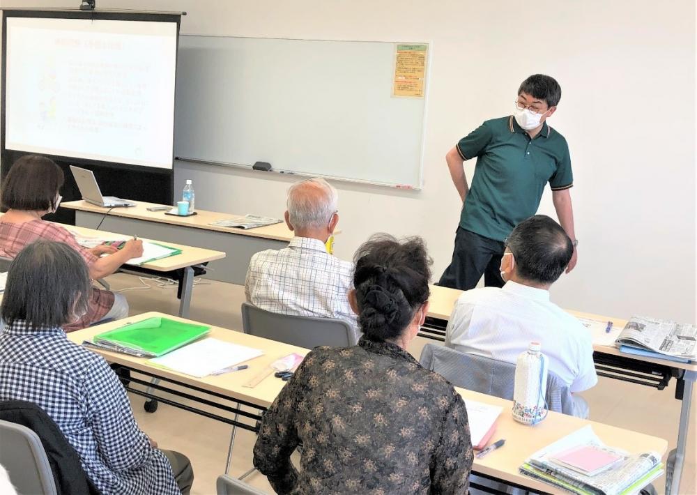 「1日1動！リアル体験教室」で講師が参加者に説明している様子。参加者は高齢者で、教室内で学びの場を共有している。
