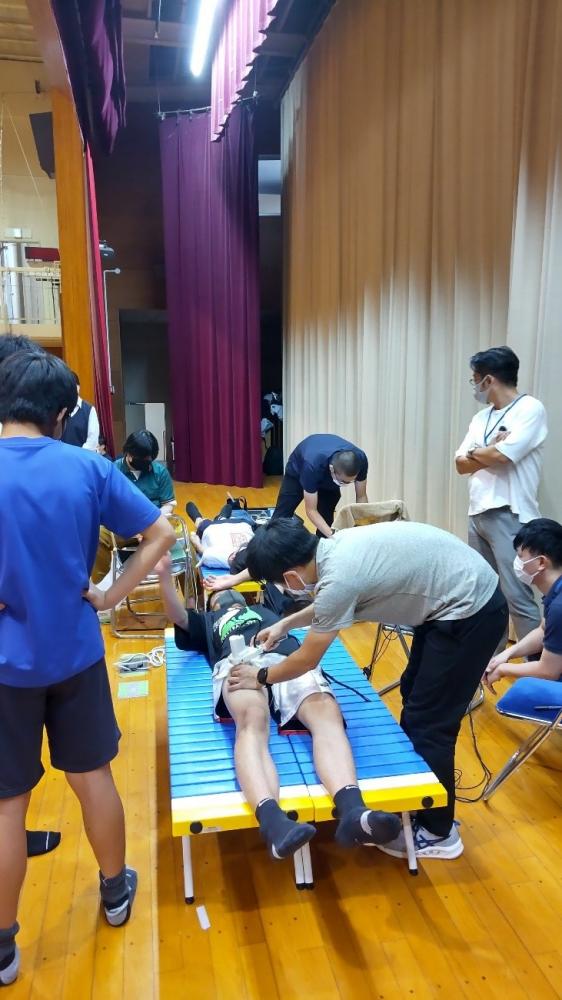 高校の体育館で、筋疲労測定を行う学生たちの様子。測定台に横たわる生徒と、周囲でサポートする教員や学生。