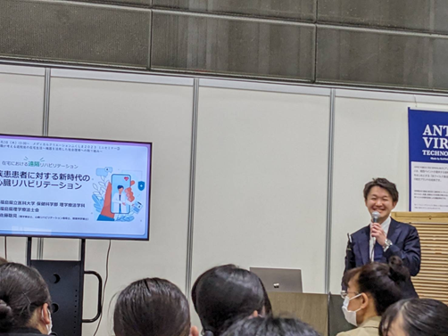 理学療法学科の教員が講演を行い、聴衆に向けて話している様子。背景にはプレゼンテーション用のスクリーンがある。