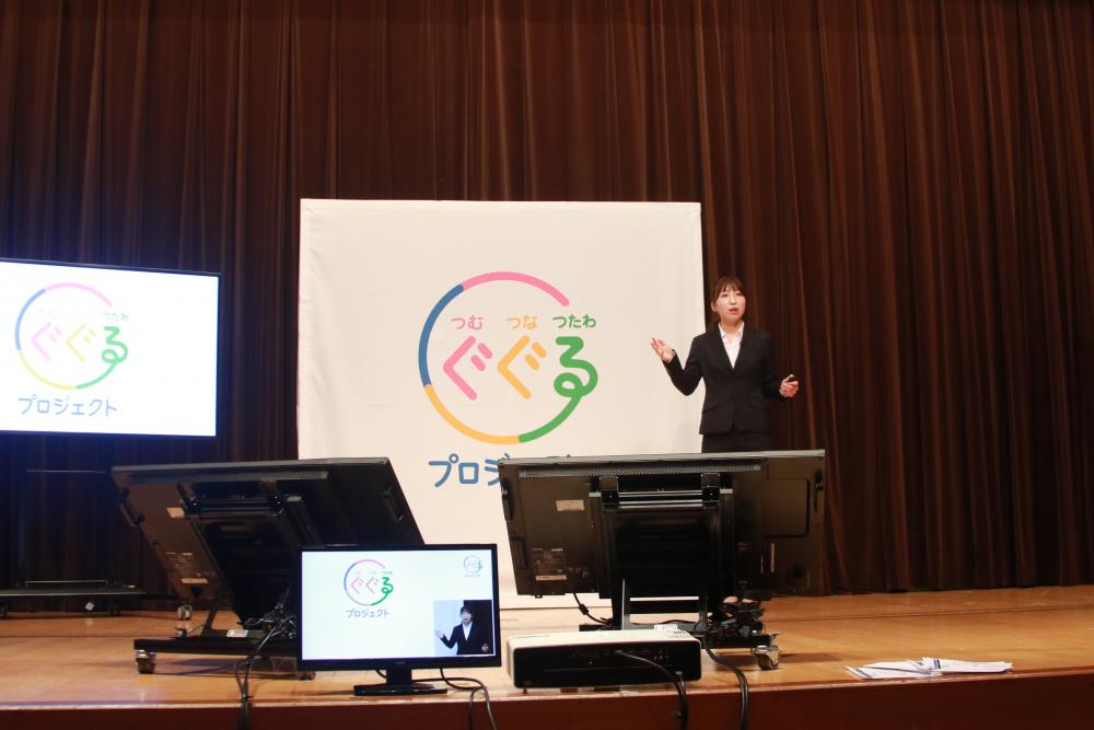 大学生が「ぐぐるプロジェクト」の発表会でプレゼンテーションを行っている様子。背景にはプロジェクトのロゴが掲示されている。