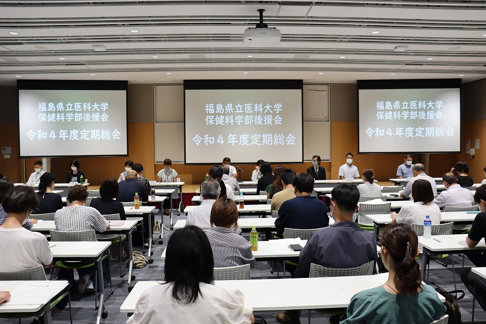 福島県立医科大学で開催された保健科学部後援会の定期総会の様子。参加者が集まり、スクリーンに議題が表示されています。