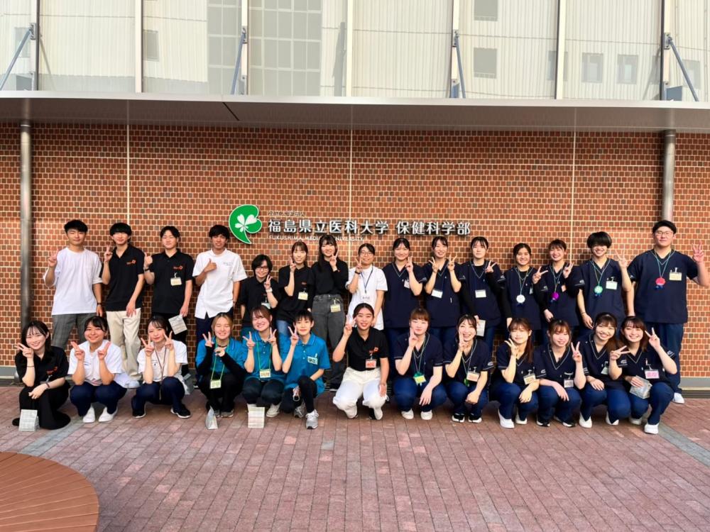 福島県立医科大学の前でボランティアサークルのメンバーが集合し、笑顔でポーズをとっている写真。