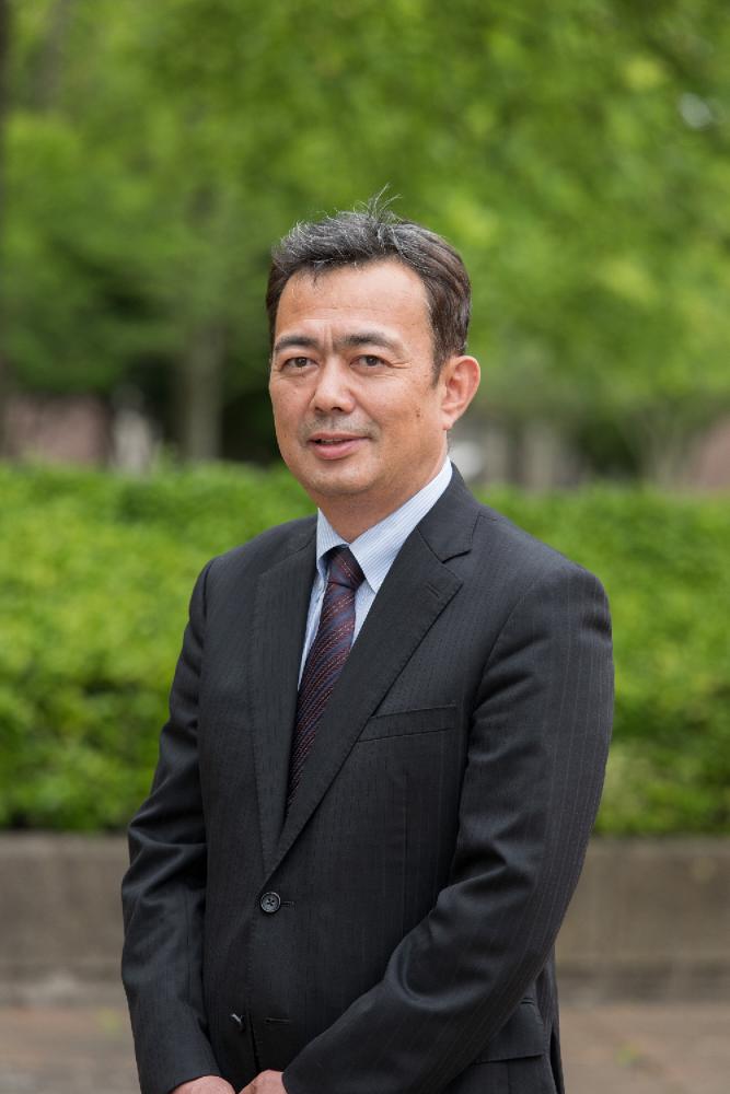 福島県立医科大学の新医療系学部設置準備室長がスーツ姿で微笑む写真。背景には緑豊かな木々が見える。