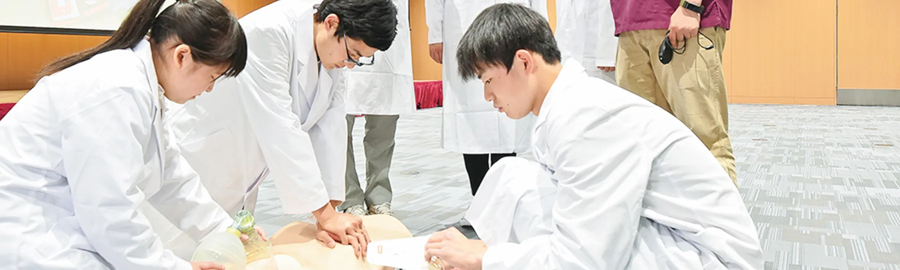 医療実習中の学生たちが心肺蘇生法を学ぶ様子。白衣を着た学生が人形に対して手を置いている。