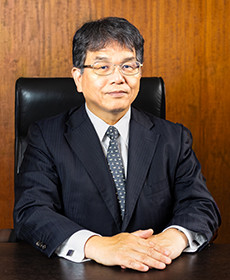 医学部長の伊関 憲氏が椅子に座り、真剣な表情でカメラを見つめているポートレート。