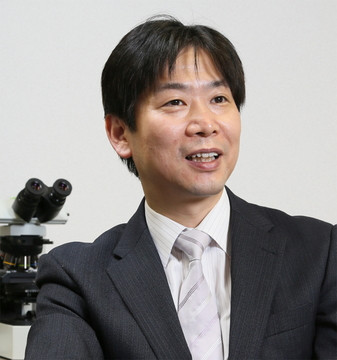 教授が教室で挨拶をしている様子。背景には顕微鏡が置かれ、研究に対する熱意が感じられる。