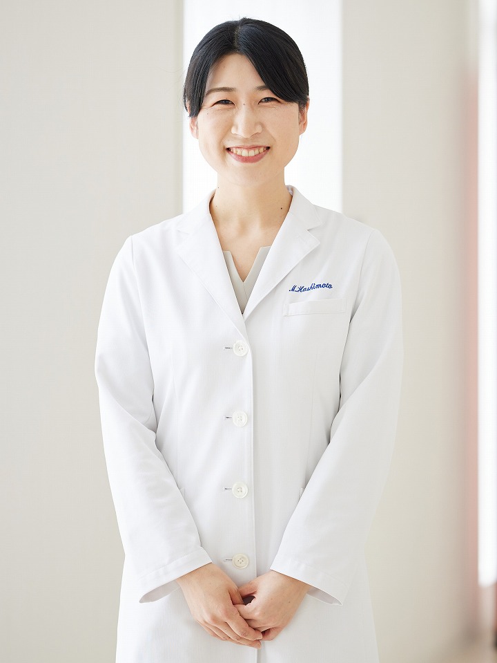 福島県立医科大学の助手、橋本万理さんのプロフィール写真。白衣を着て青い背景の前で微笑んでいる。