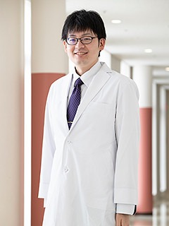 病院助手の阿部貞彦さんが白衣を着て微笑んでいる姿。福島県立医科大学の医療センター内で撮影された写真。