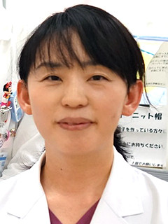 病院助手の吉田清香さんが白衣を着て微笑んでいる写真。背景には医療関連のポスターが見える。