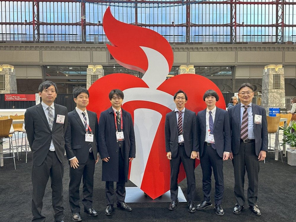 6人のスーツ姿の男性が、炎と心臓をデザインした大きな赤いオブジェの前で並んで立っています。背景にはガラス窓があり、開放的な空間が広がっています。