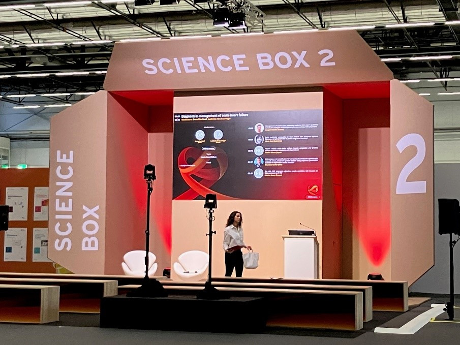 白いブラウスを着た女性が「SCIENCE BOX 2」と書かれたステージ上でプレゼンテーションの準備をしている。スクリーンには心不全に関する情報が映し出されている。