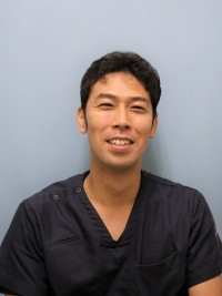 スタッフ紹介の写真で、笑顔の佐藤彰彦講師が医療用の服を着ている。背景は青色。