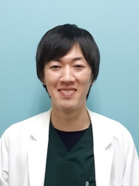 病院助手の室田定洋さんが白衣を着て微笑んでいる写真。背景は淡い青色。