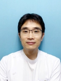 医療白衣を着た男性が青い背景の前で微笑んでいる写真。