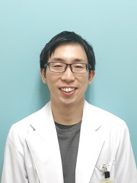 冨田湧介が白衣を着て微笑んでいる写真。背景は淡い青色。