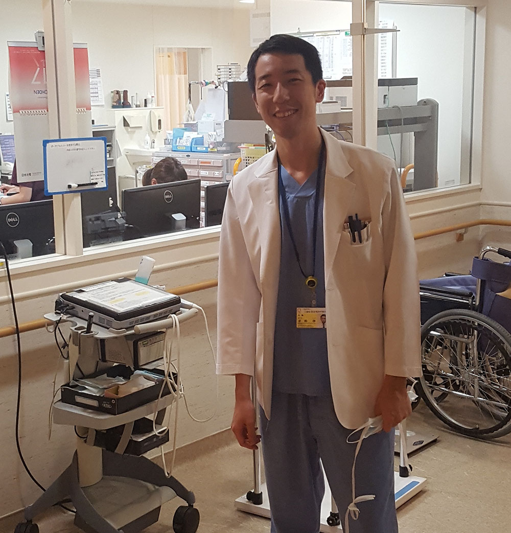 心臓血管研究所付属病院で白衣を着た医師が笑顔で立っている様子。背景には医療機器や車椅子が見える。