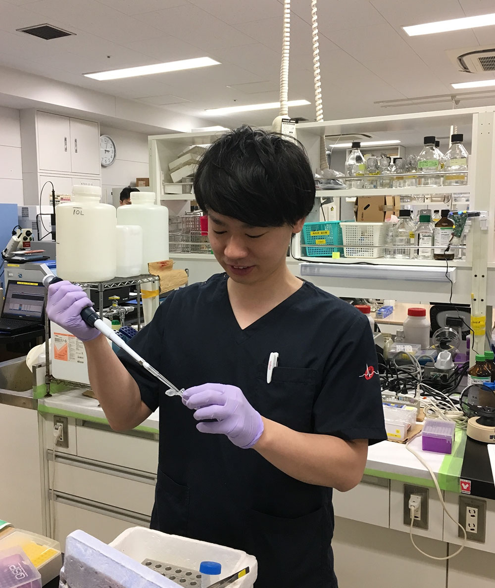 研究室で実験を行う黒い服を着た男性がピペットを使用している。背景には実験器具が並んでいる。