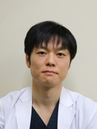 医療白衣を着た男性がカメラに向かって微笑んでいるポートレート。背景はシンプルな壁。