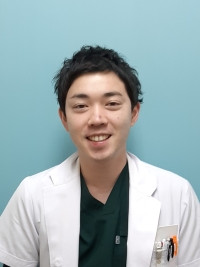 医療現場で働く若い医師が白衣を着て微笑んでいる写真。背景は淡い青色。