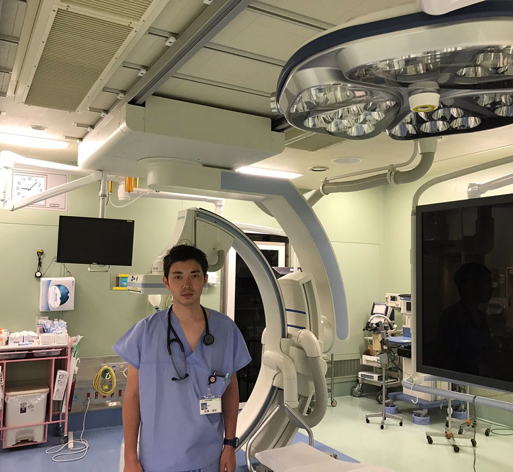 医療現場で青いスクラブを着た男性が立っている様子。手術室の機器や照明が背景に見える。