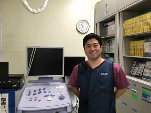 国立循環器病研究センターの医師が、医療機器の前で微笑んでいる写真。背景には書類棚と時計が見える。