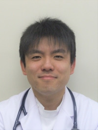 白衣と聴診器を身に着けた医師が笑顔で前を見ている。背景は白で、医療に関連するプロフェッショナルな印象。