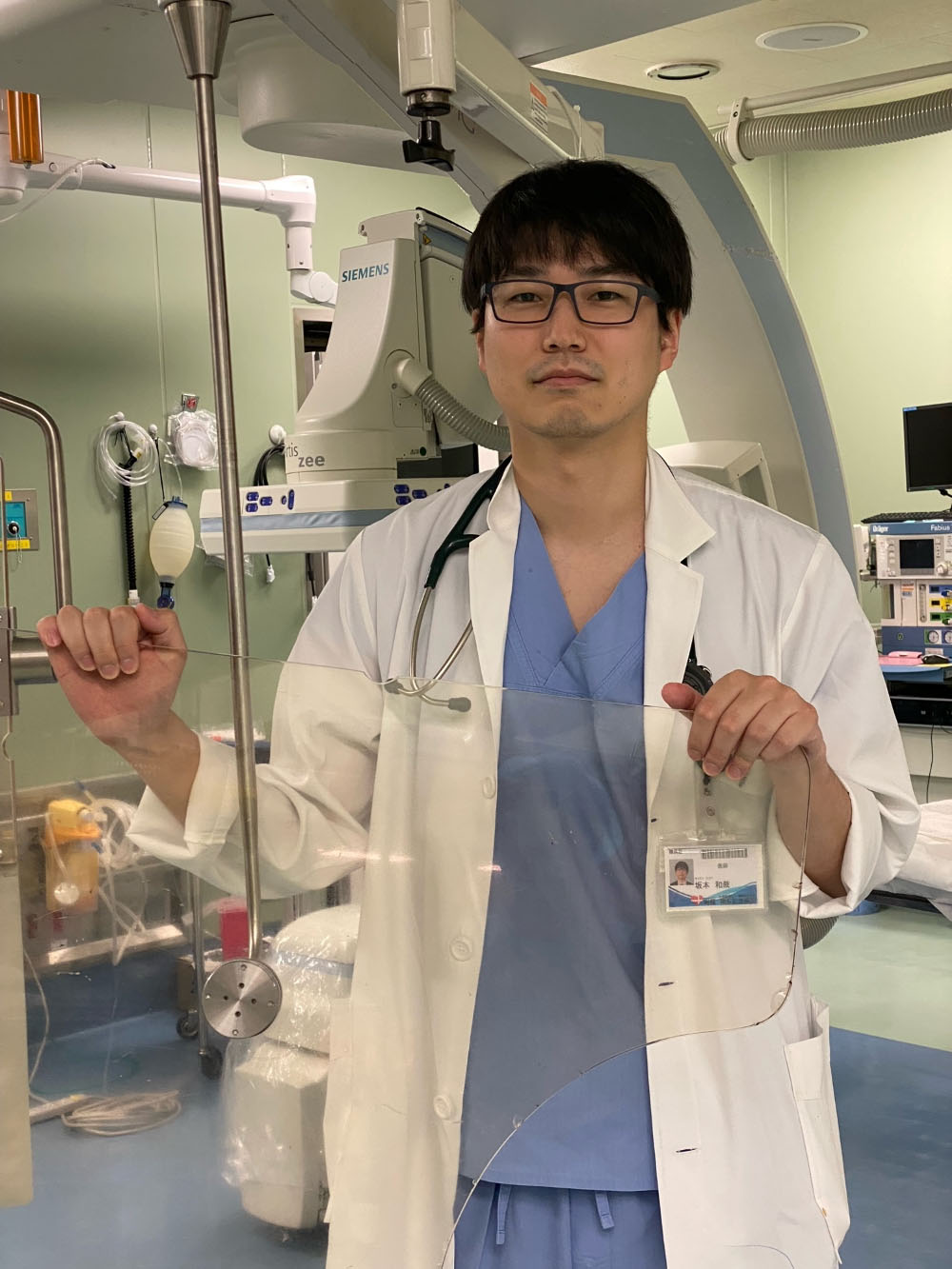 医療現場で白衣を着た男性が、手に器具を持ちながらカメラに向かって立っている様子。