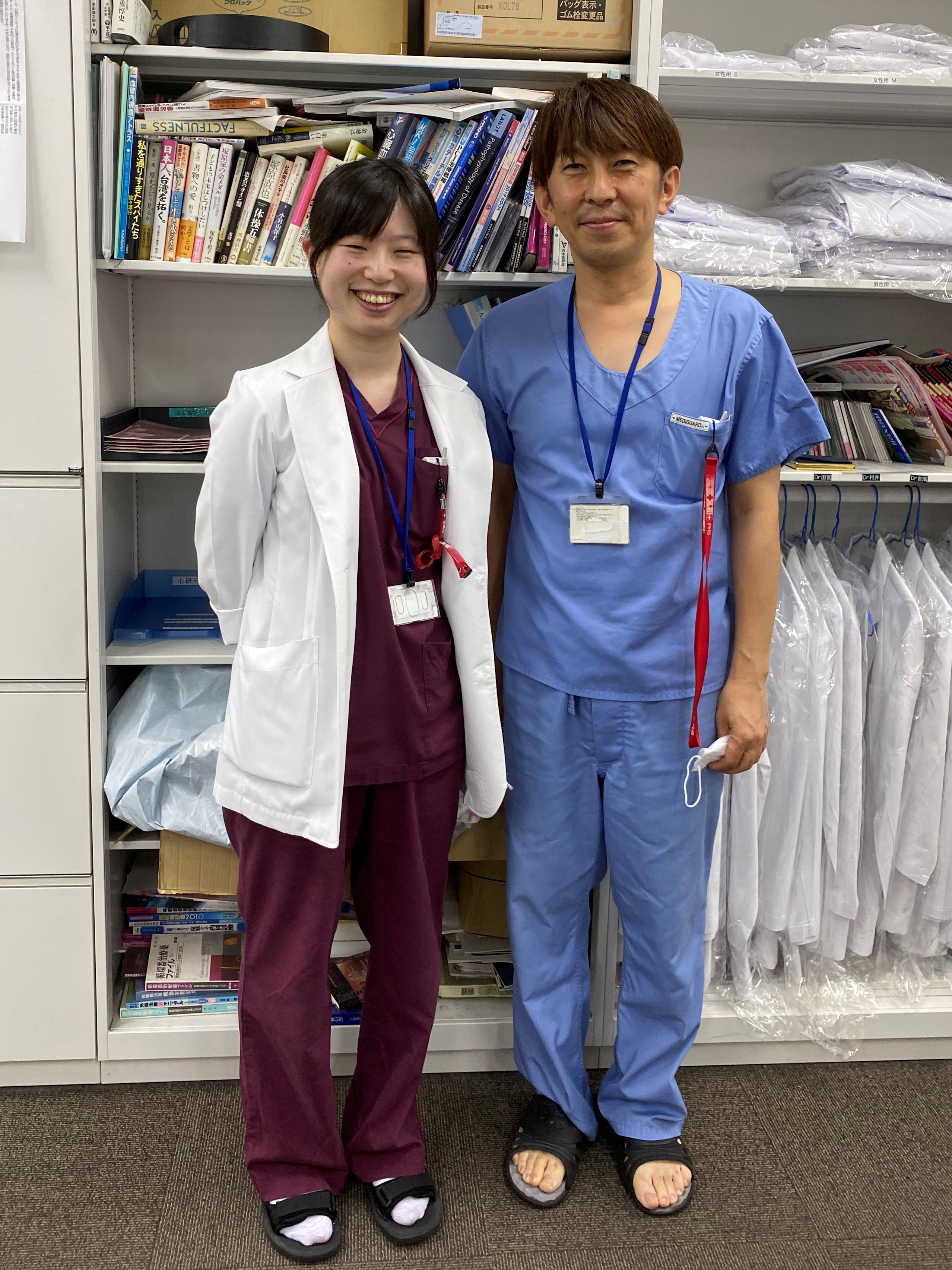 医療現場で白衣を着た女性と青いスクラブを着た男性が並んで立っている写真。背景には医療用の道具や衣類が見える。