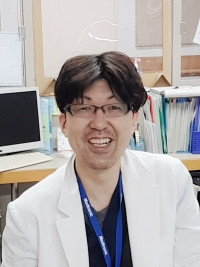 医療現場で白衣を着た佐藤崇匡さんが笑顔でカメラを見つめている写真。背景には書類やパソコンが見える。