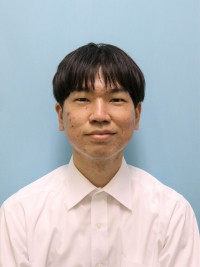 大学院生の笠原慎也さんが白いシャツを着て、青い背景の前で微笑んでいるポートレート。