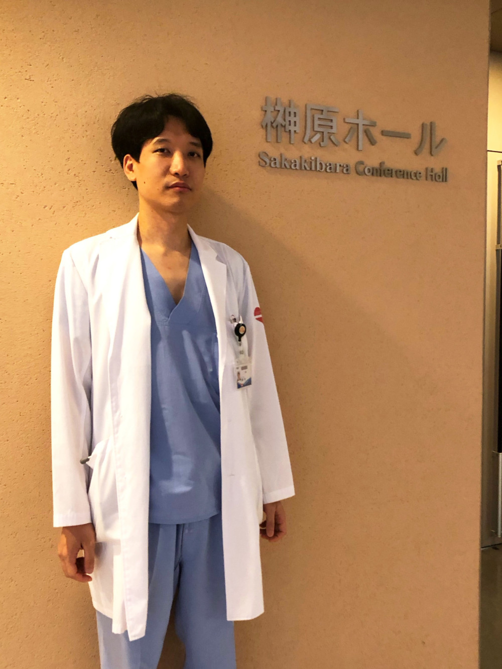 白衣を着た男性が榊原ホールのサインの前で立っている。背景はクリーム色の壁。