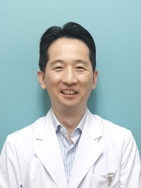 医療白衣を着た男性が青い背景の前で微笑んでいる写真。