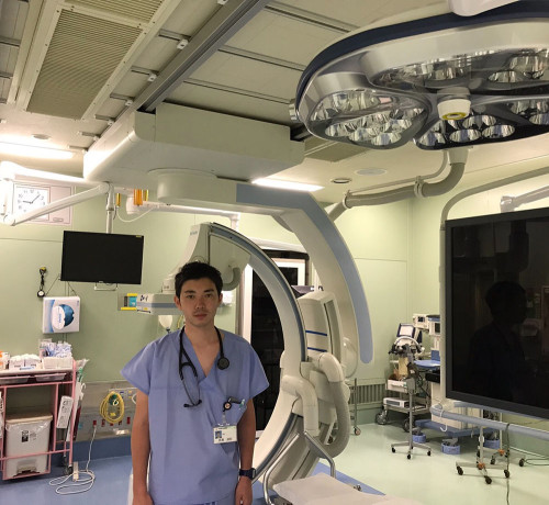 医療現場で青いスクラブを着た男性が立っている様子。手術室の機器や照明が背景に見える。