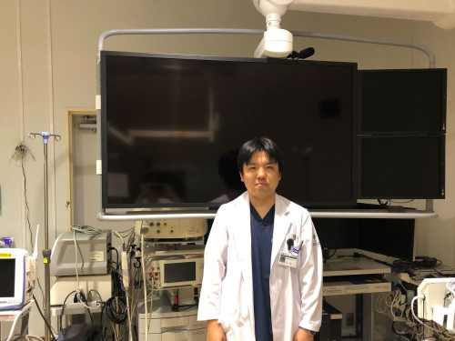 医療機器の前で白衣を着た医師が立っている病院の内装。大型モニターと様々な医療機器が背景に見える。