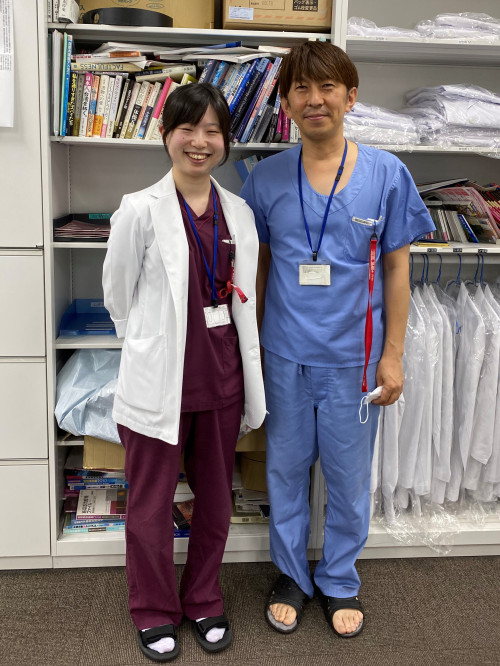 医療現場で白衣を着た女性と青いスクラブを着た男性が並んで立っている写真。背景には医療用の道具や衣類が見える。
