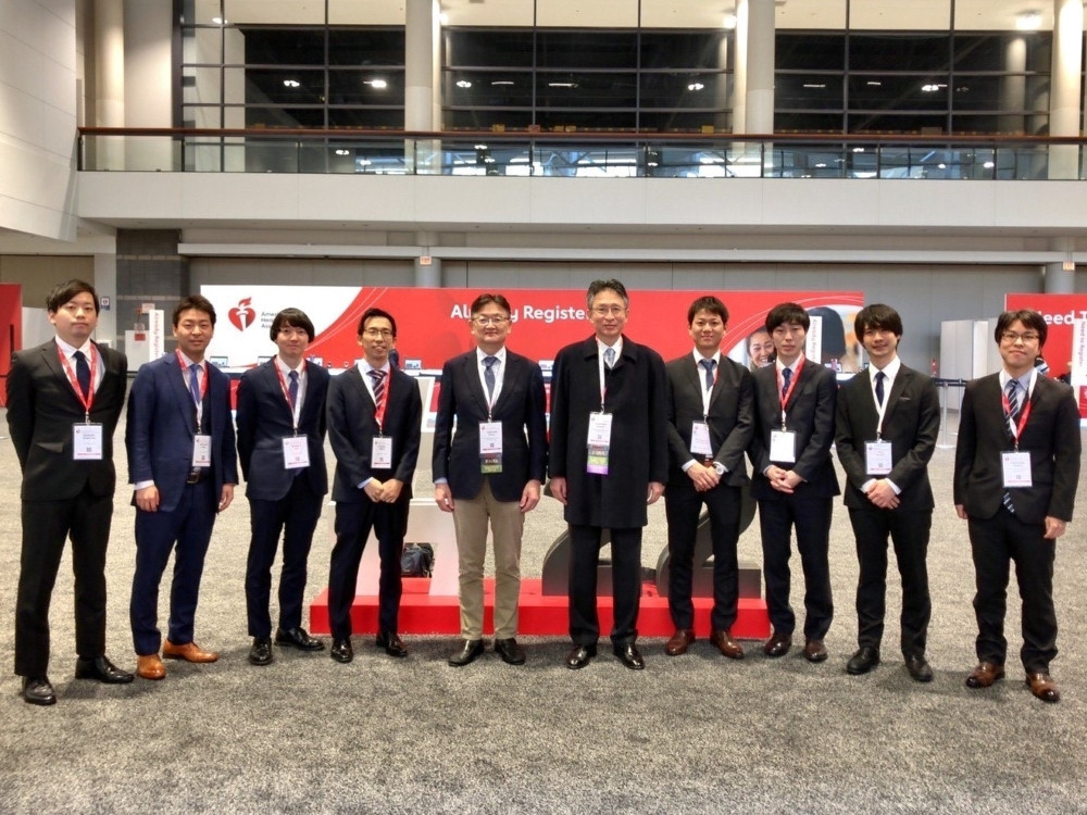 スーツを着た男性たちが展示会場で集合写真を撮影している。背景には登録カウンターと赤い装飾が見える。